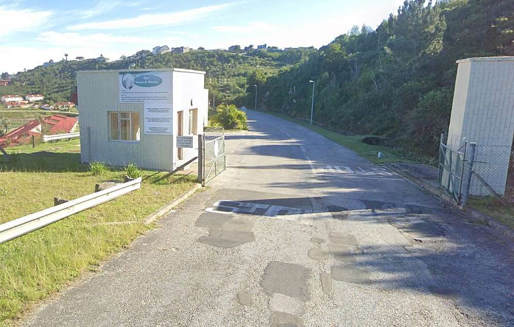 The potholed patched grey tar road leads through the open gate of the prison entrance road. To the left is the unmanned guard house.