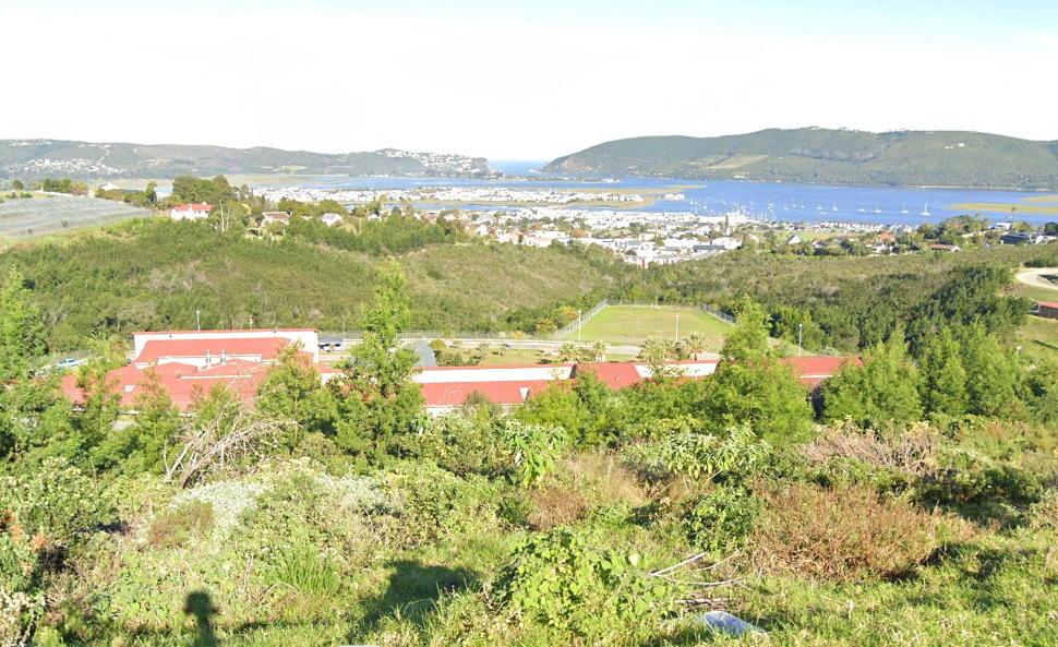 In the foreground the red roof of the Prison is seen while above and beyond the roof of the building, the town of Knysna sprawls in the distance towards the twin cliffs of the Knysna Heads.