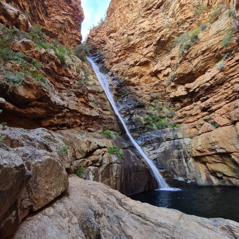 A sliver stream of water cascades down a narrow slot of the river as the waterfall drops forty metres from the break in the towering sandstone clifs, to fall into deep clear water pool below. Small green shrubs are seen to line the sheer sides of the cliffs.