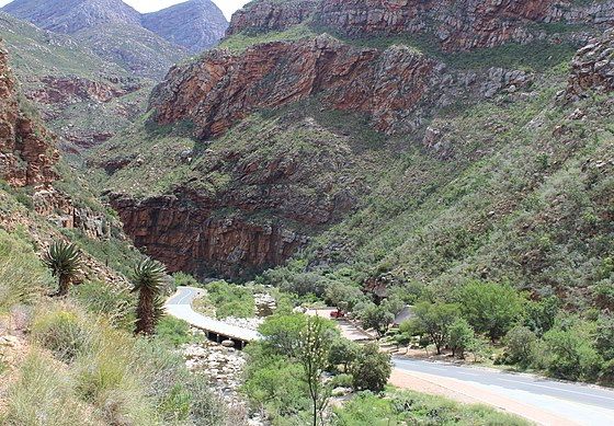 A two lane road winds its way across a dry riverbed. High towering sandstone cliffs with green bushes and shrubs on each side of the road. The river drops through a series of waterfalls.