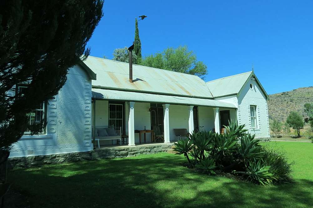 Cool lush green lawn grass leads up to a Victorian style farm house painted in white with a steel roof. Here and there patches of light rust are to be seen on the roof. Couches and wooden tables are on the patio or stoep, as it is called in the Karoo.