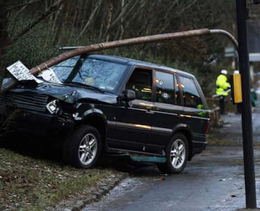 A dark-coloured Range Rover stands busted against a lamppost that is bent over the car. The back window of the car show two star shaped shattering with a hole in the middle of each. Clearly bullet holes.