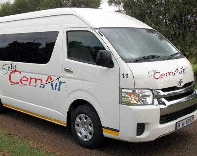 A white sixteen seat mini bus with the red and blue logo of CemAir, driving past on the gravel road.