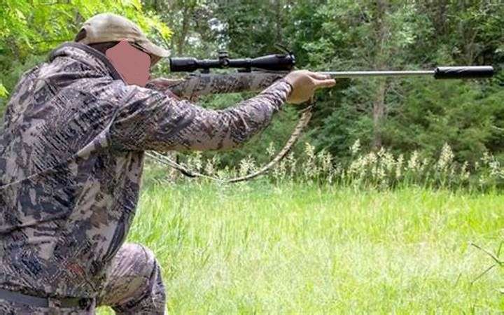 A unidentified person in camo clothing sighting a hunting rifle. He/she is completely surrounded by low shrubs and bush.