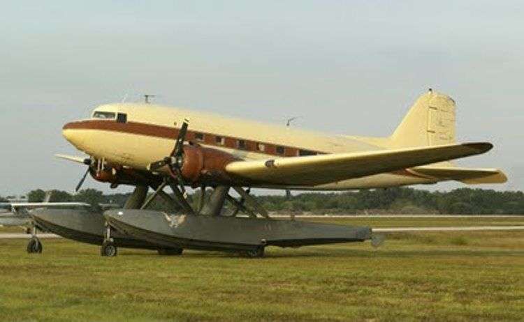 A cream-yellow DC-3 aircraft with light brown accents sits high on huge pontoon floats. The four wheels under the floats visible in the picture.