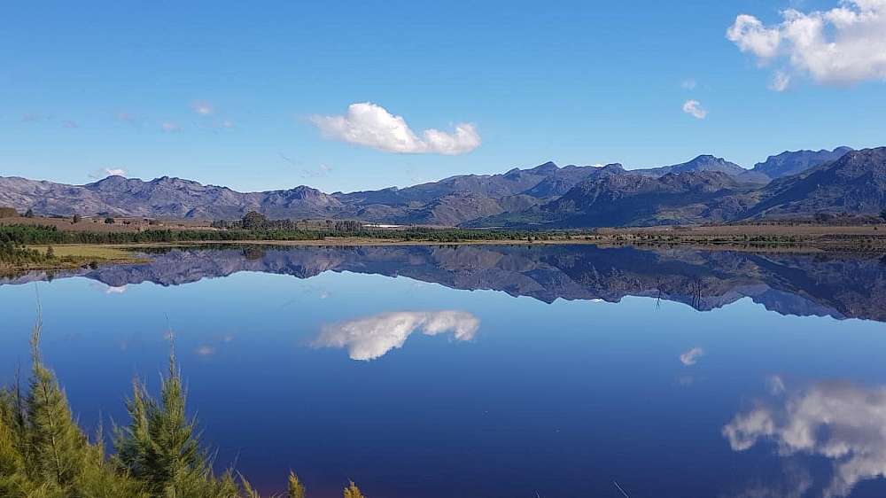 The white foggy clouds reflect of the glass calm waters of the Teewaterskloof Dam. The blue jagged ridge mountain of the Hottentots Holland Mountains reflect off the surface of the waters, almost like a mirror. The far bank of the dam is a picture of green trees and cultivated farm land.