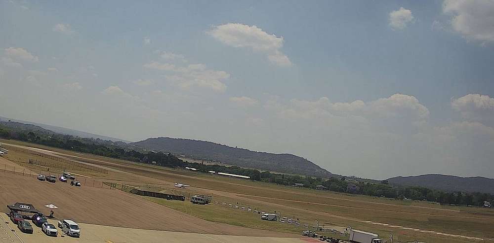 The view from the terrace of the Villa San Giovanni’s Ristorante over the A apron of Wonderboom airport. In the foreground vehicles are seen servicing aircraft. The view cuts across Runway 11/29 towards the north with two hills on the horizon.