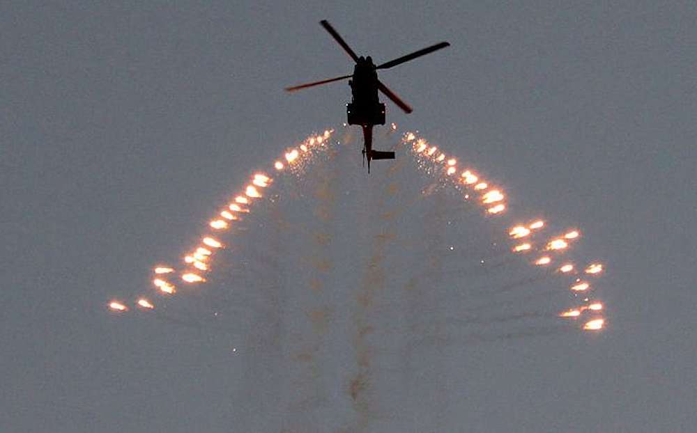 The blue sky is darkened to a dull grey as the Oryx helicopter fires a stream of bright orange and white infra-red decoy flares from its left and right counter measures tubes. The bright luminance of the flares dropping away from the helicopter in an attempt to distract any missiles fired towards it.