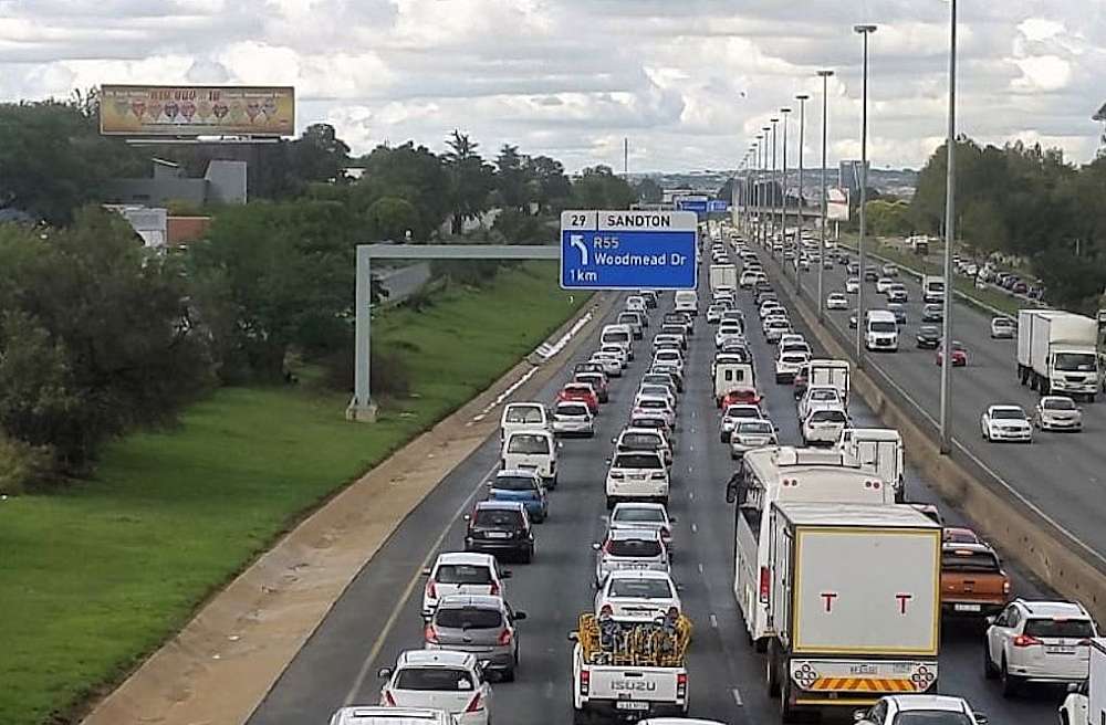 A picture showing the south bound traffic on the N1 Highway, Midrand, Johannesburg, Gauteng, South Africa. The sky is slightly overcast and along the four-lane southbound N1 track green grass and medium sized trees are seen. An overhead traffic sign show that the highway is in the suburb of Sandton with the next turn-off to be Woodmead Drive, one kilometre away.