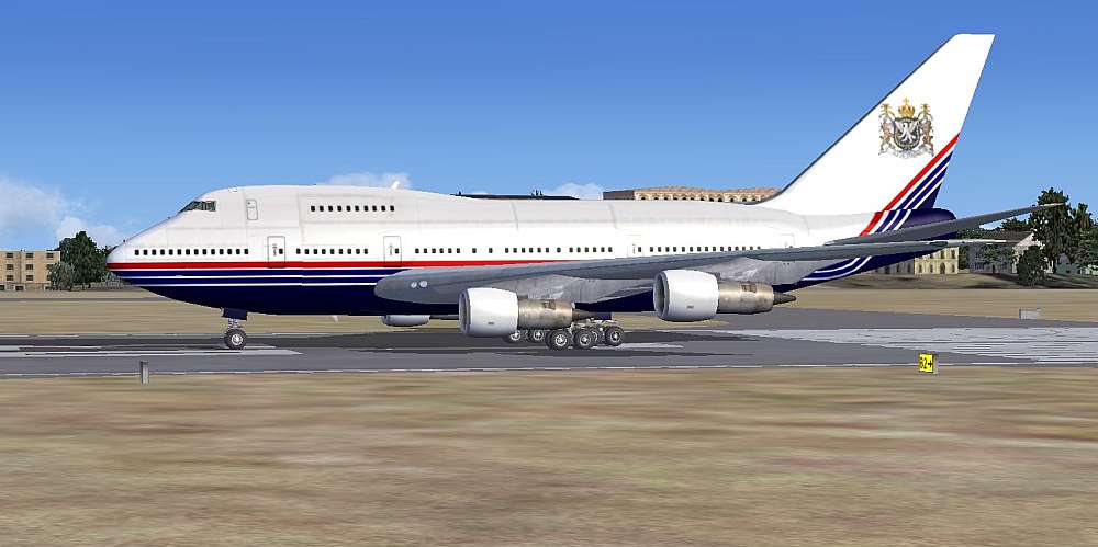 A side view of the Queen’s flight Boeing 747SP waiting on the apron at George Airport. The aircraft is overall white on the top with a royal blue underside. One red and two royal blue stripes flash along the fuselage and strike up onto the tail past the Royal Crest of the Queen of Volynia.
