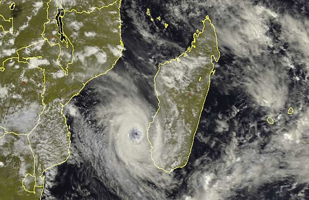 A high-altitude satellite weather map shows a powerful tropical cyclone centered in the Mozambique Channel, positioned directly between the western coast of Madagascar and the eastern coast of Mozambique. The cyclone exhibits a classic spiral structure with a distinct, clear ‘eye’ at its center, surrounded by a dense, white swirl of clouds indicating intense wind speeds. Heavy cloud cover stretches outward from the storm, beginning to drape over the southern tip of Madagascar and moving toward the African mainland. The landmasses of Southeast Africa are highlighted with yellow borders, providing a clear sense of the storm’s proximity to the coast and its inevitable path toward the interior.
