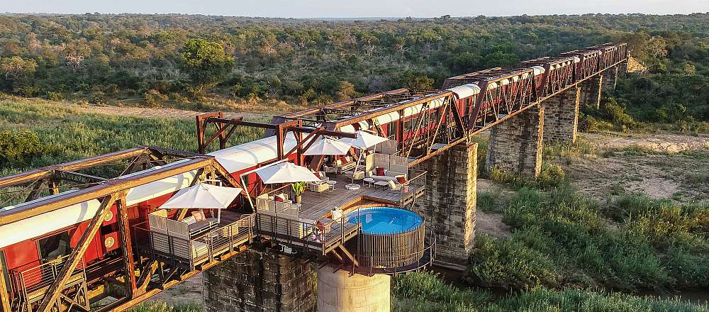 A breathtaking wide-angle, high-resolution photograph of the famous Kruger Shalati ‘Train on a Bridge’ hotel. A line of vintage-style luxury train carriages is permanently stationed across the historic Selati Bridge over the Sabie River. The scene is bathed in the golden light of late afternoon. One carriage features a stunning overhanging wooden deck with white umbrellas, lounge seating, and a circular, blue-tended plunge pool suspended directly over the riverbed far below. The vast, green canopy of the Kruger National Park stretches to the horizon under a clear sky, highlighting the unique blend of industrial history and modern luxury.