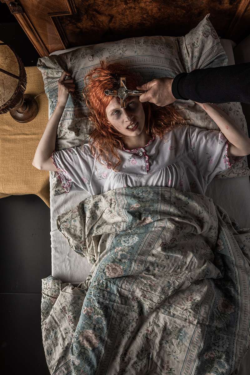 Color photo of an angry redheaded woman lying in bed staring up at a preist performing an exorcism with no visible pupils...