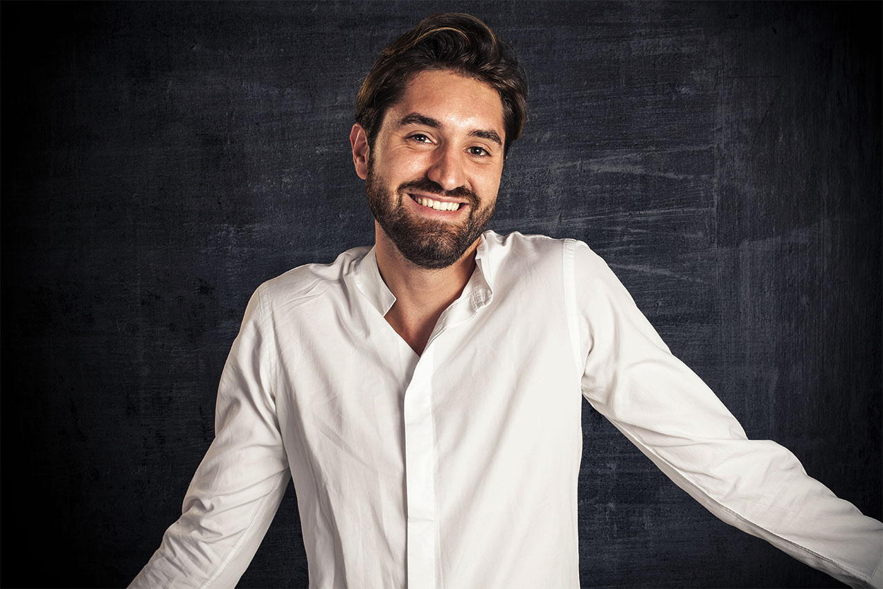Color photo of a shy bearded man in an open shirt shrugging, with a wry grin standing before a classroom blackboard.