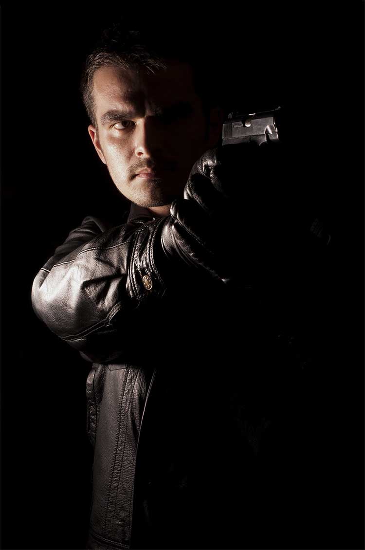 Photo of an armed man in a heavy leather jacket aiming a pistol at someone on a black background.