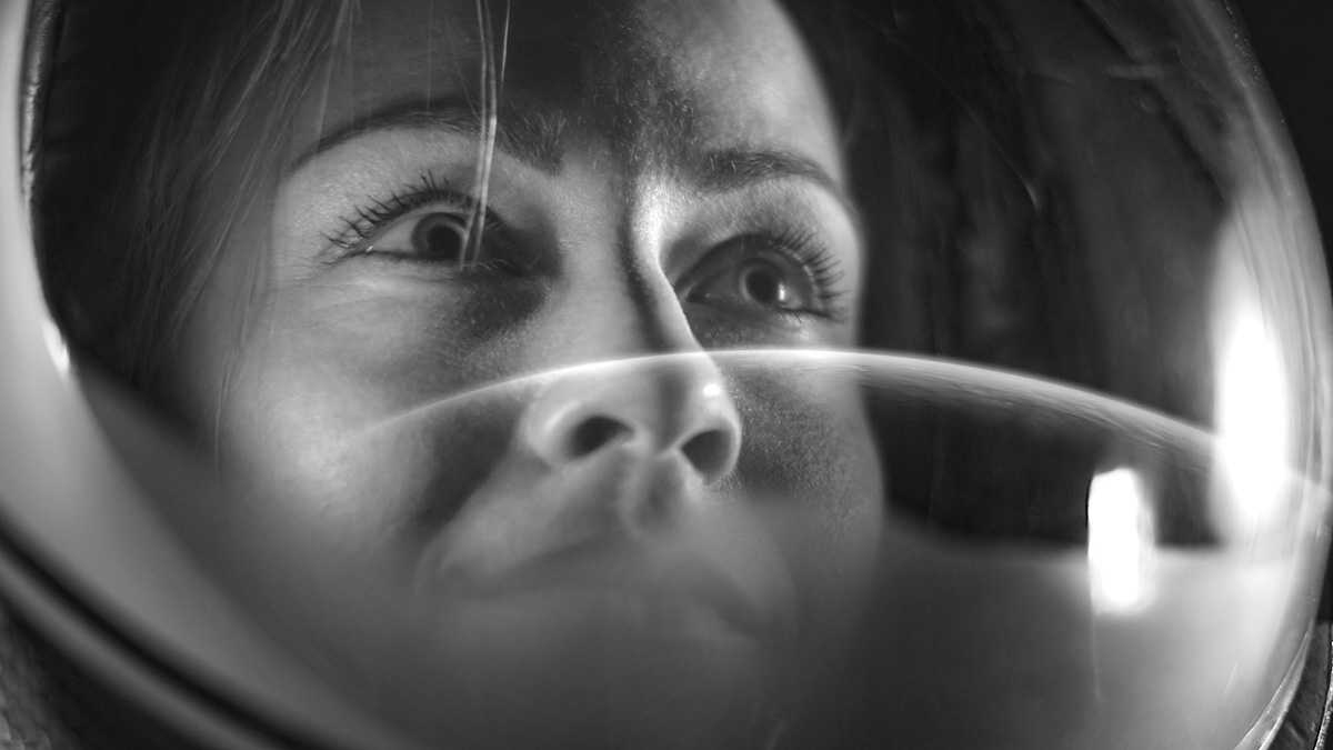 Black and white image of a female human as seen through her helmet faceplate as she watches her new home from space.